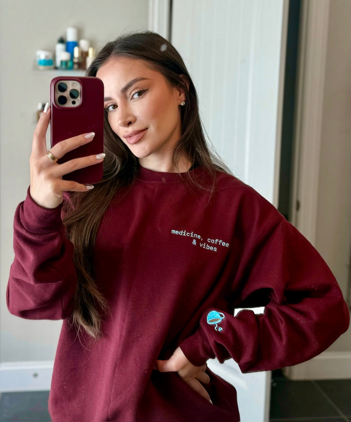 Dr Daisy wearing a maroon Medicine, Coffee & Vibes sweatshirt with coffee cup graphic on sleeve, taking a mirror selfie.