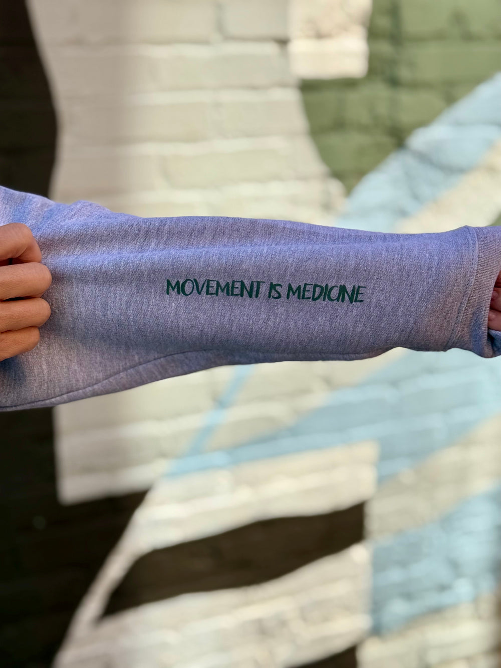 Person wearing a gray sweatshirt showing off the left sleeve text: 'MOVEMENT IS MEDICINE', and standing against a blurred background.