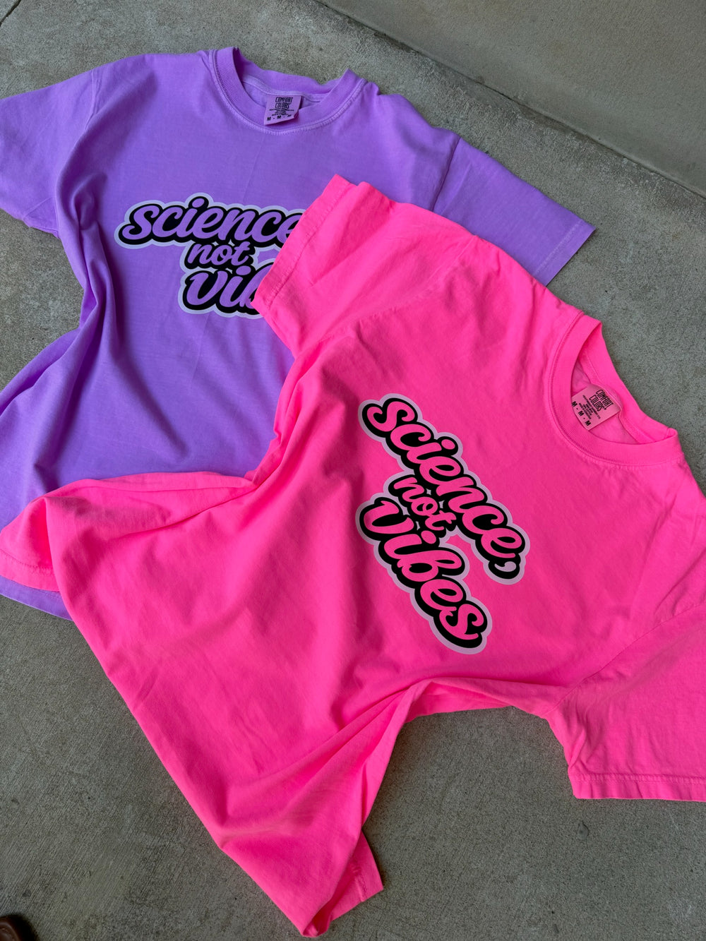 Purple and pink t-shirts with 'Science, not vibes' text on a concrete surface