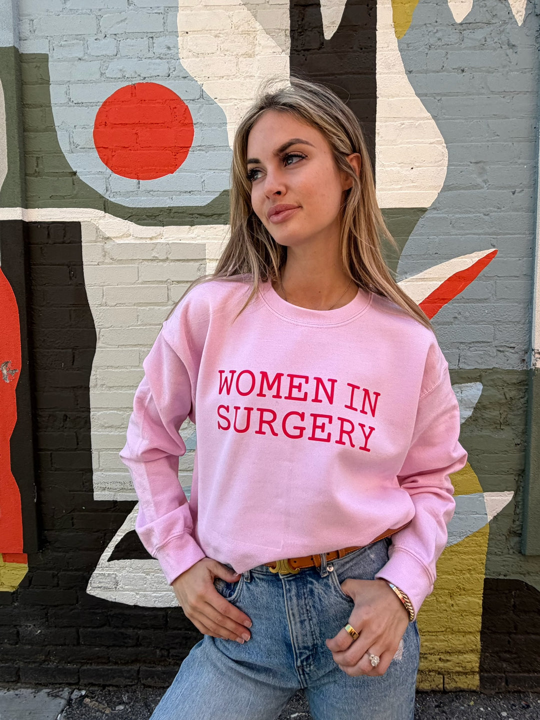 Woman wearing a pink sweatshirt with 'WOMEN IN SURGERY' text, standing in front of a colorful mural.