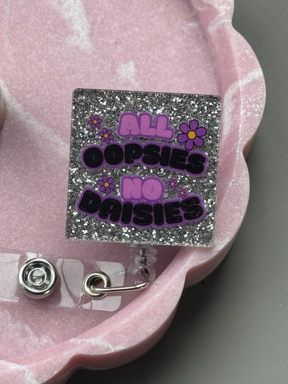 Retractable nurse badge reel with silver glittery background and bold text: “All Oopsies No Daisies” in purple and black with flower accents displayed in pink tray.