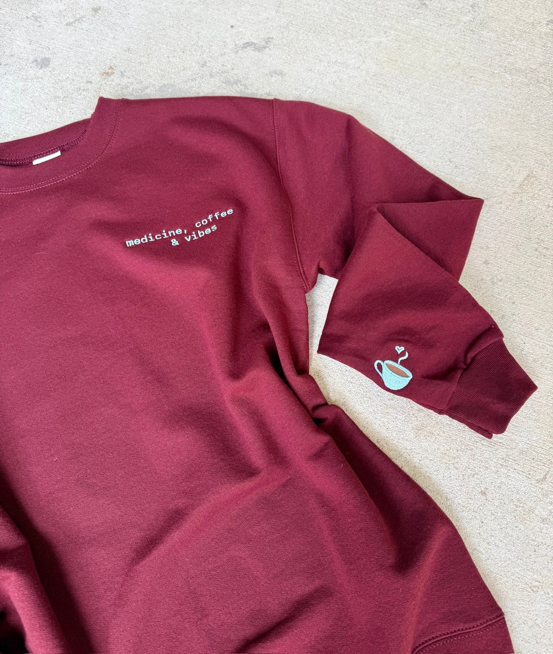 Maroon sweatshirt with medicine coffee & viebs and coffee cup graphic on a light gray surface