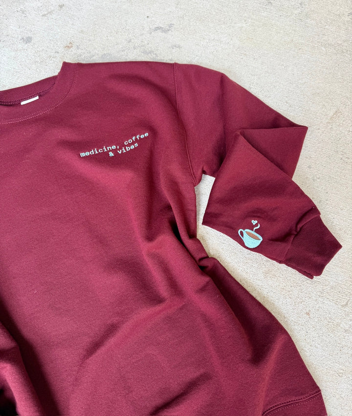 Maroon sweatshirt with medicine coffee & viebs and coffee cup graphic on a light gray surface