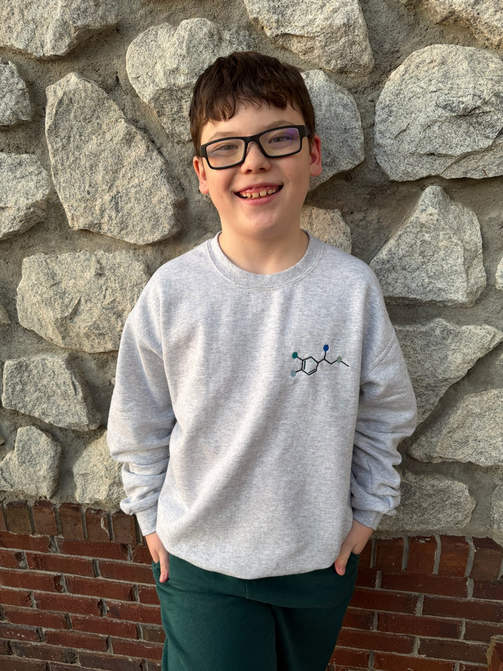 Child wearing a gray sweatshirt with an embroidered Epinephrine (Adrenaline) Molecule design against a stone wall.