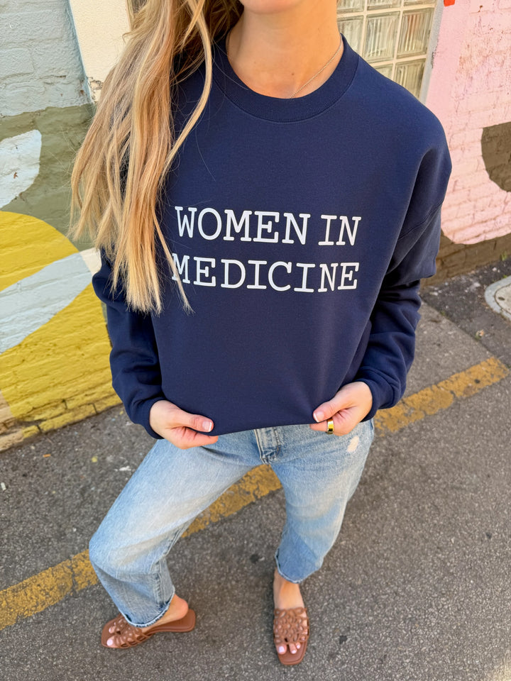 Person wearing a navy sweatshirt with white 'WOMEN IN MEDICINE' text, standing on a street.
