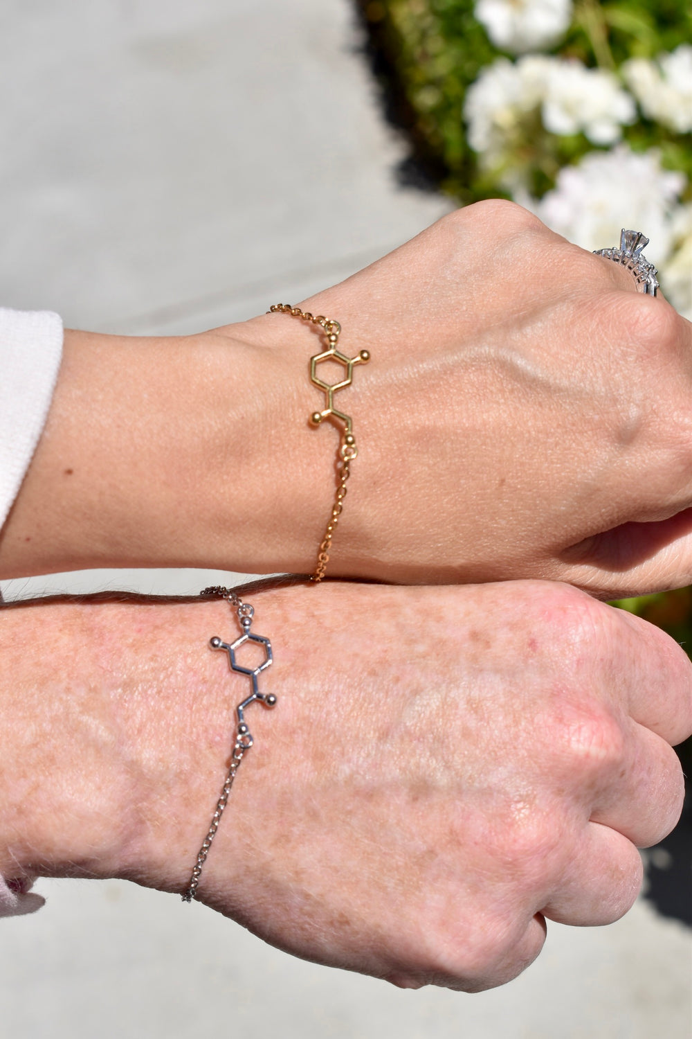 Two hands wearing noradrenaline molecular design bracelets with a blurred floral background