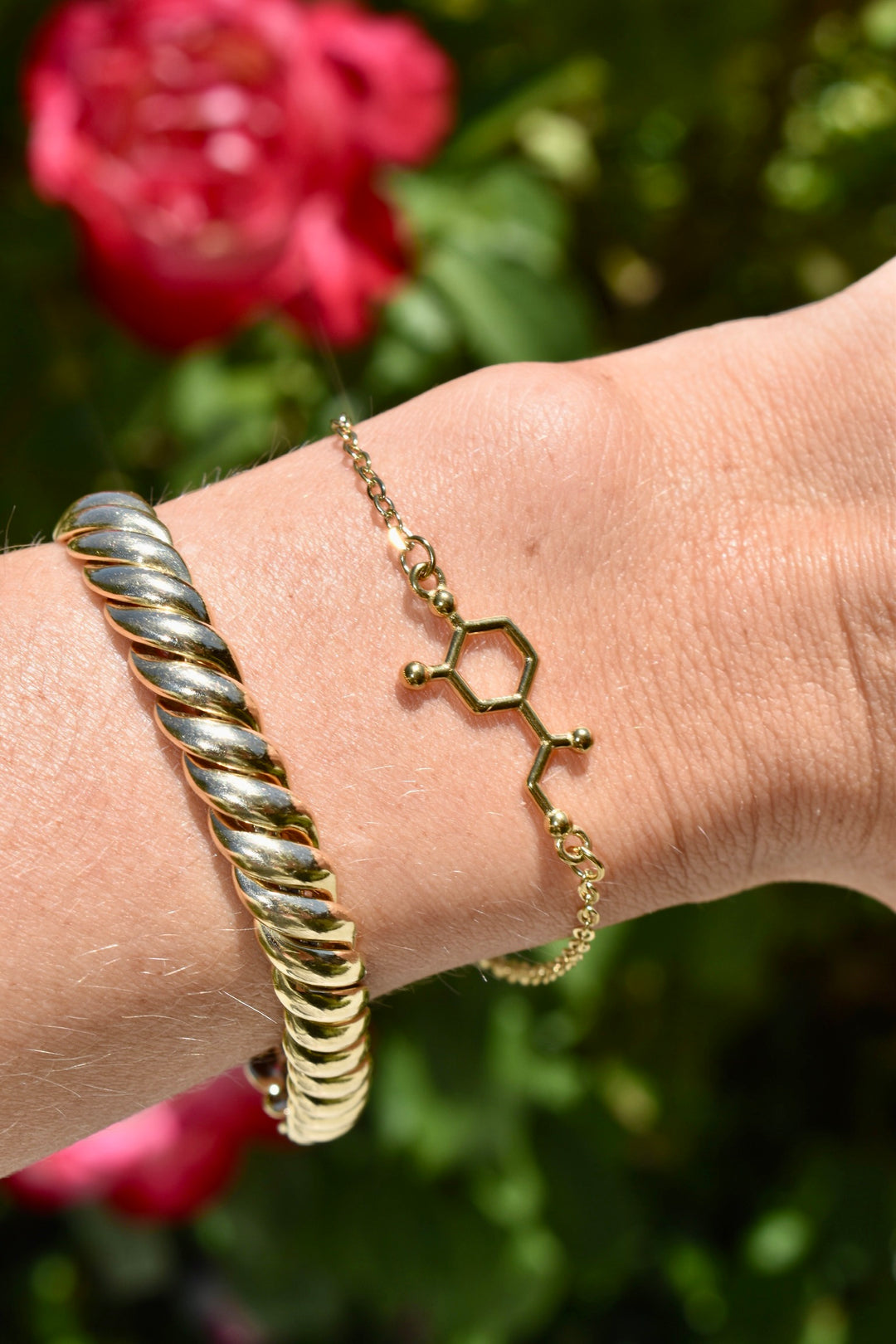 Gold noradrenaline molecule bracelet with a gold rope bracelet on a wrist with a blurred floral background