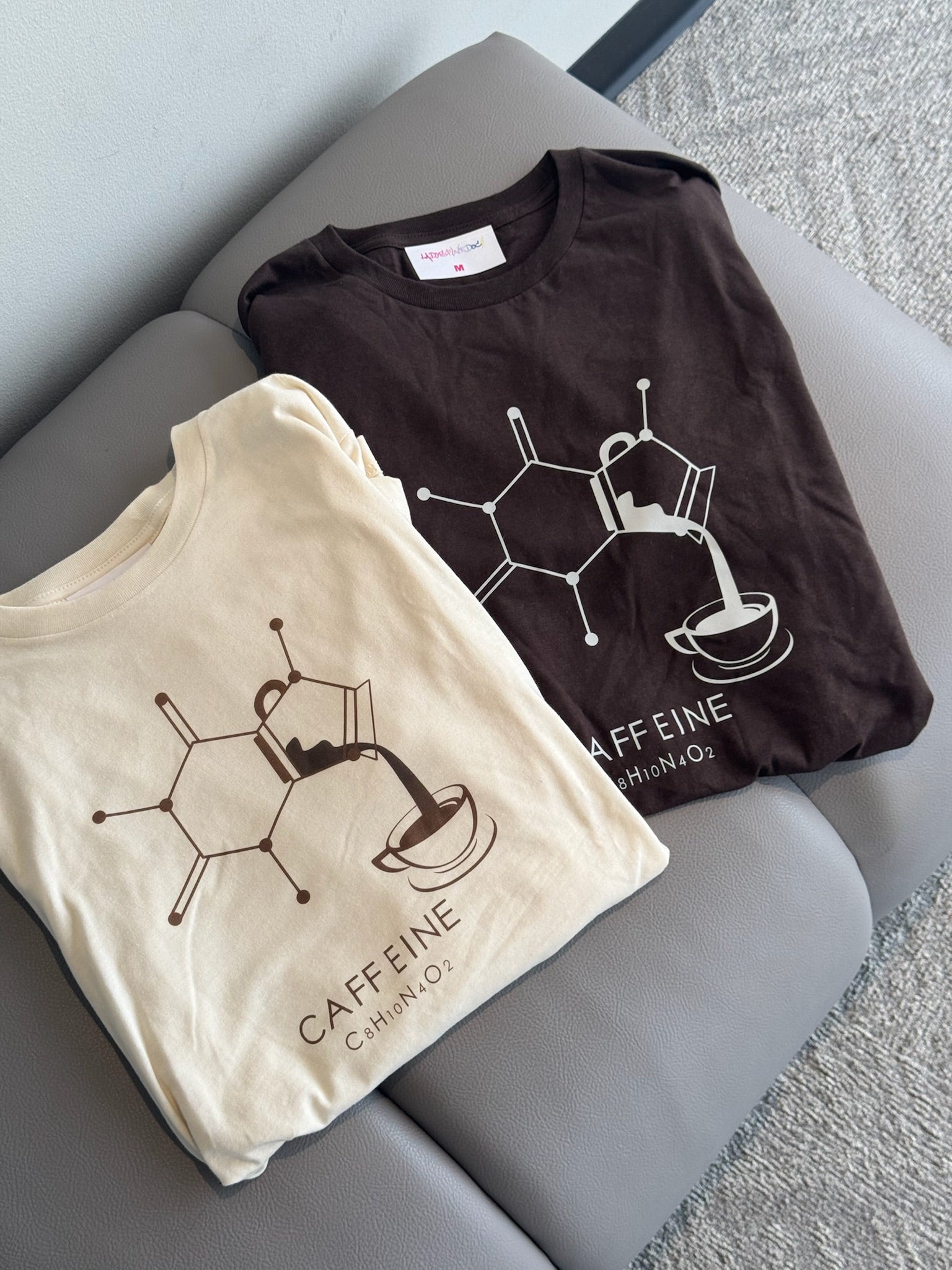 Two folded t-shirts, one brown, the other cream, with a coffee molecule design on a gray bench.