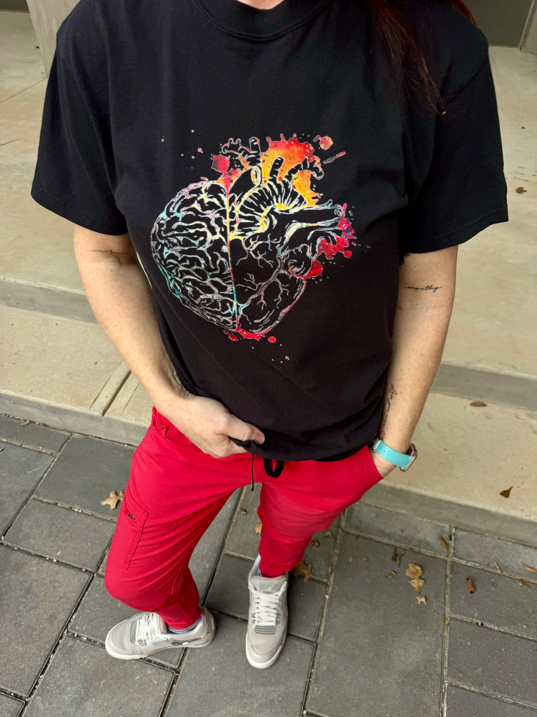 Ladyspinedoc wearing a black t-shirt with a colorful, anatomical brain/heart graphic design, standing on a sidewalk.