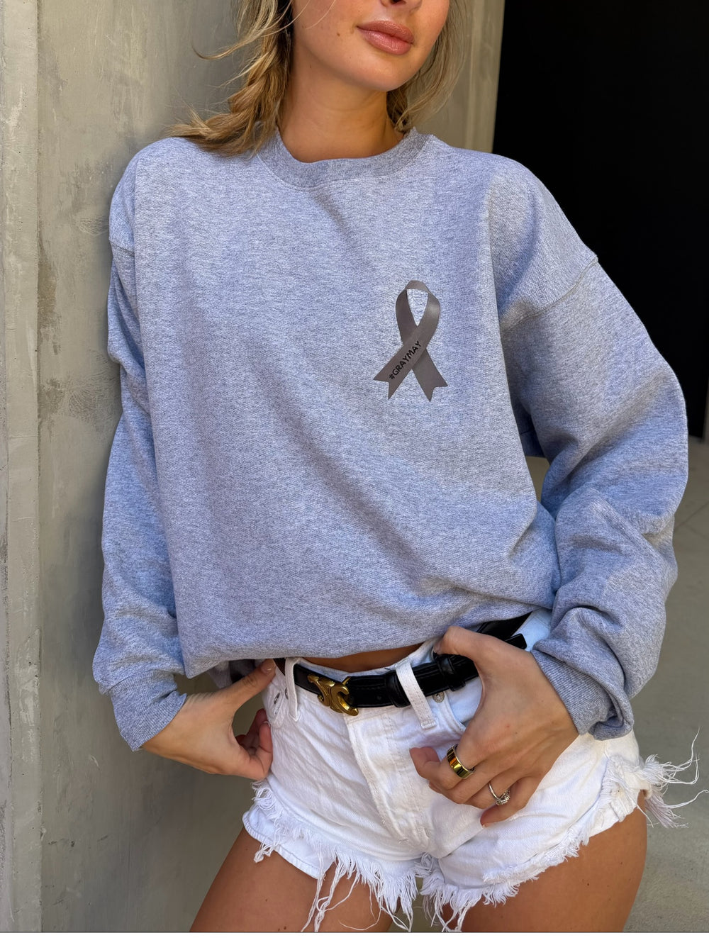 Person wearing a gray sweatshirt with a gray ribbon design, paired with white shorts. The ribbon say #graymay.