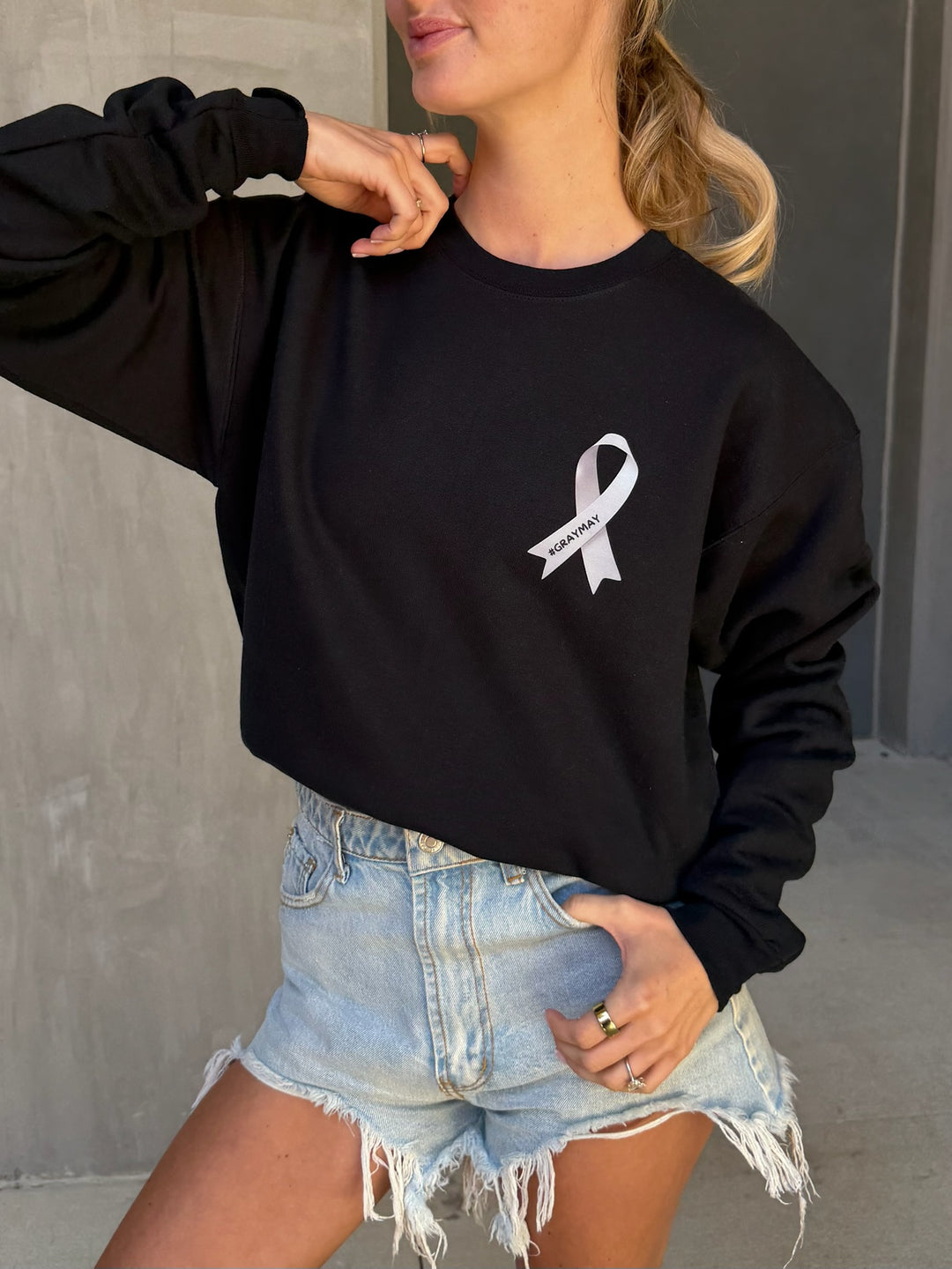 Person wearing a black sweatshirt with a gray ribbon showing #GRAYMAY logo and denim shorts.