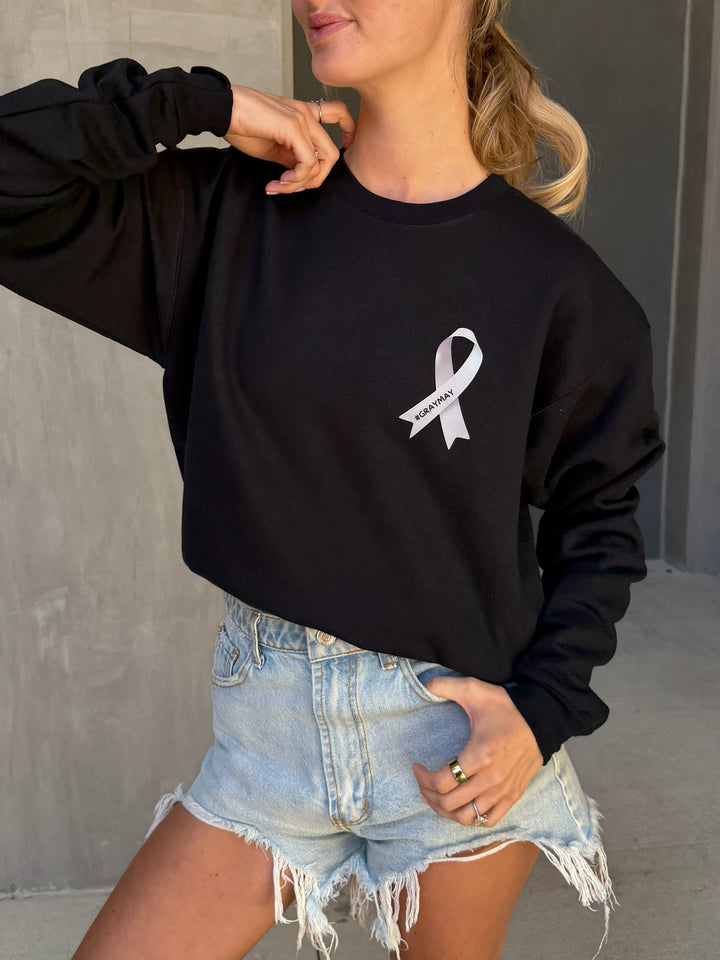 Person wearing a black sweatshirt with a gray ribbon showing #GRAYMAY logo and denim shorts.