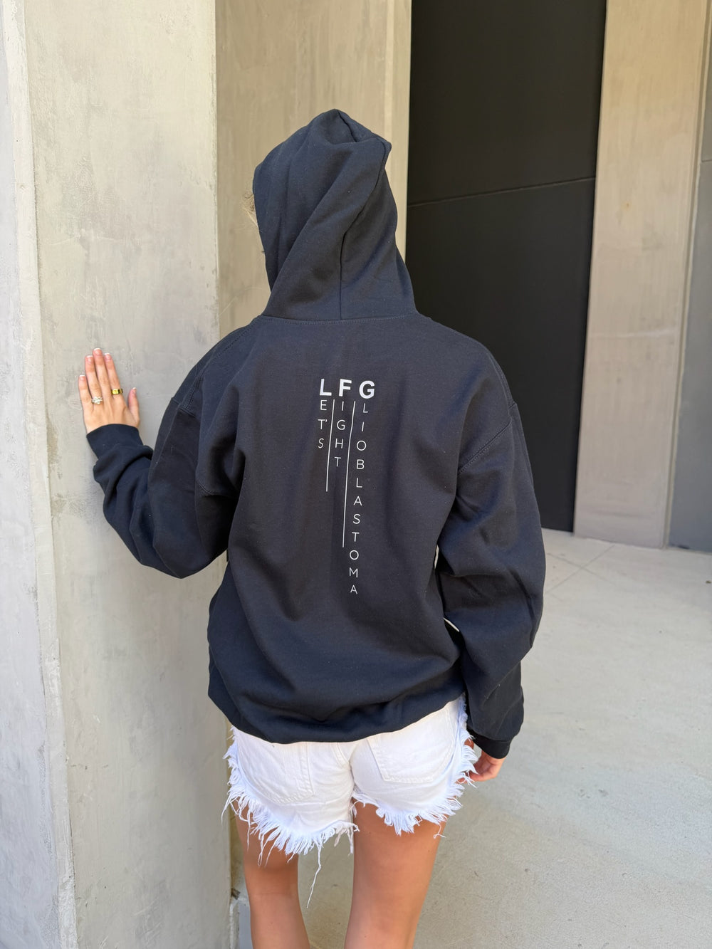 Person wearing a black hoodie with text on the back, standing against a wall.