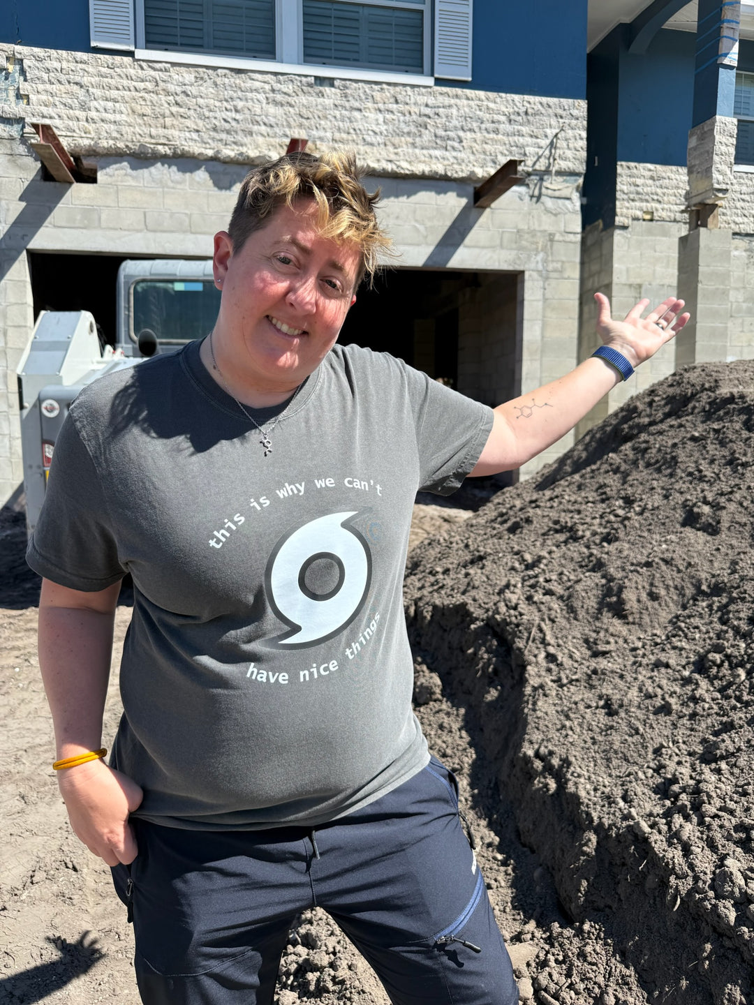Dr. Beachgem standing in front of her house near a pile of dirt and wearing a gray tee shirt that says "This is why we can't have nice things" with a hurricane symbol