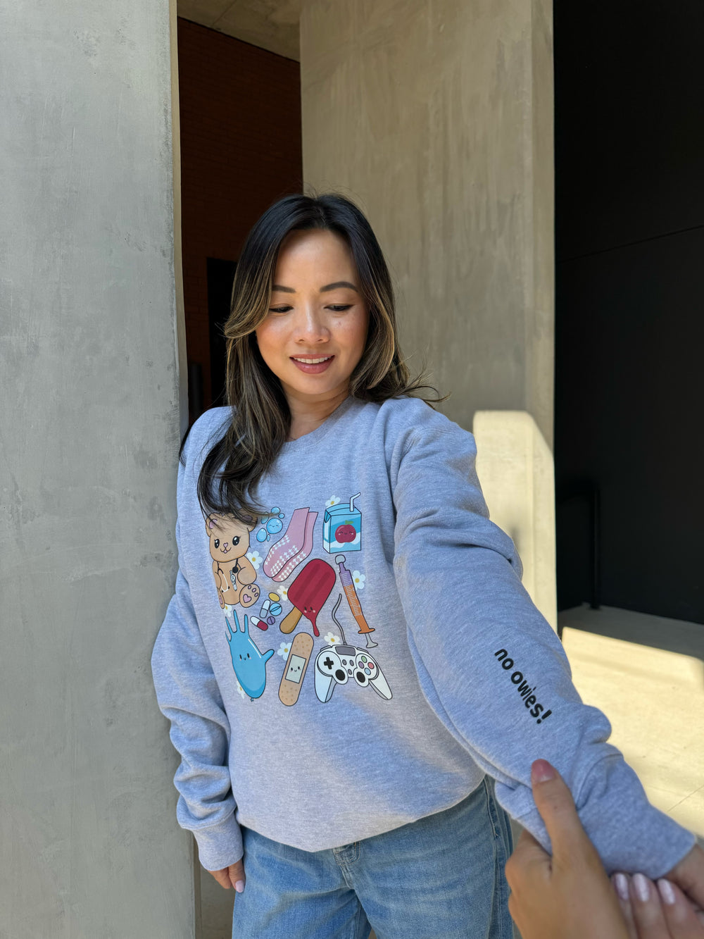 Jenn wearing the Ladyspinedoc X Scrubhacks No Owies! Pediatrics Collage Crewneck Sweatshirt - Heather Gray With Pedi Nurse Essentials and highlighting the “No Owies!” saying on Sleeve