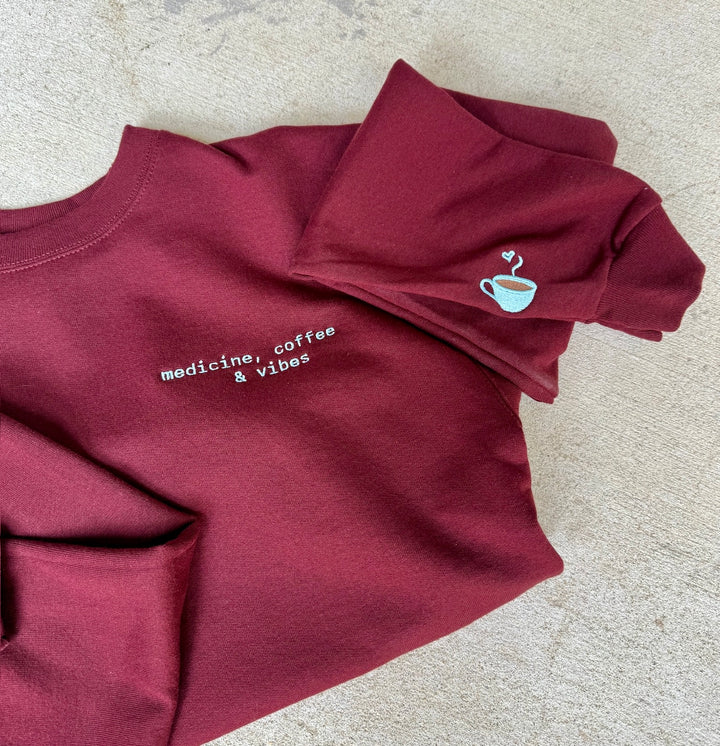 Maroon medicine, coffee, & vibes sweatshirt with coffee cup graphic on a beige surface