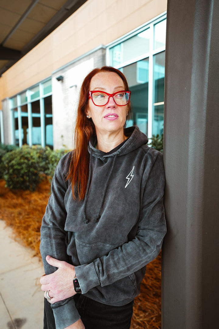 Dr. Betsy Grunch (Ladyspinedoc) facing forward wearing red glasses and the dark gray Incredible Hoodie, showcasing front lightning bolt detail.
