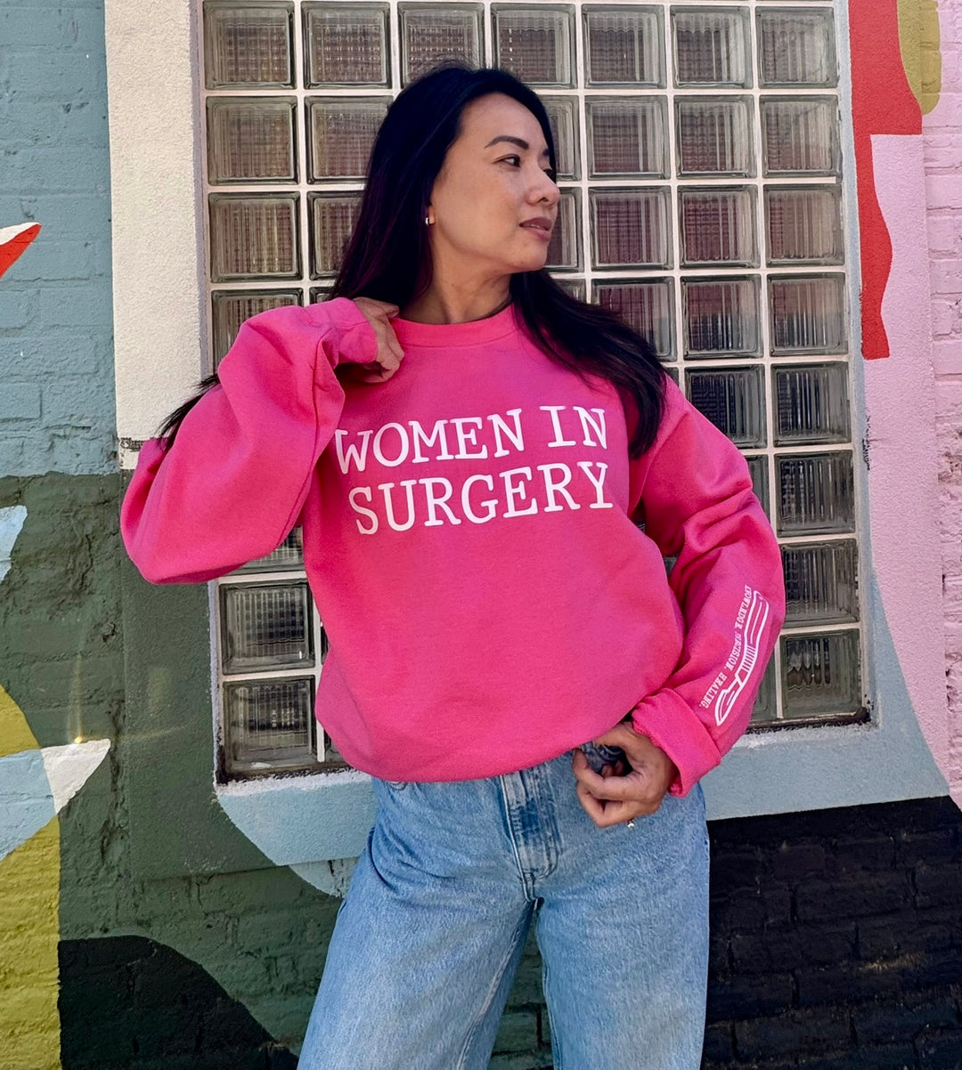 Person wearing a pink sweatshirt with 'WOMEN IN SURGERY' text, standing in front of a glass-block wall.