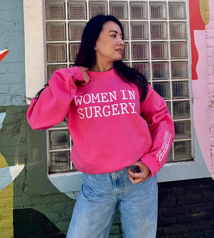 Person wearing a pink sweatshirt with 'WOMEN IN SURGERY' text, standing in front of a glass-block wall.