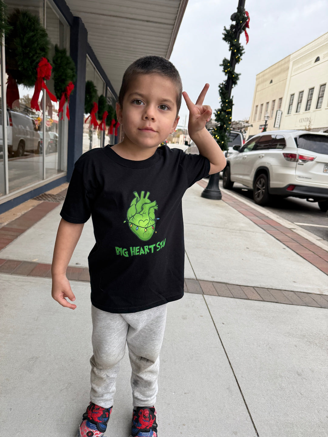 Child wearing a black Big Heart Szn t-shirt with green graphics on a sidewalk