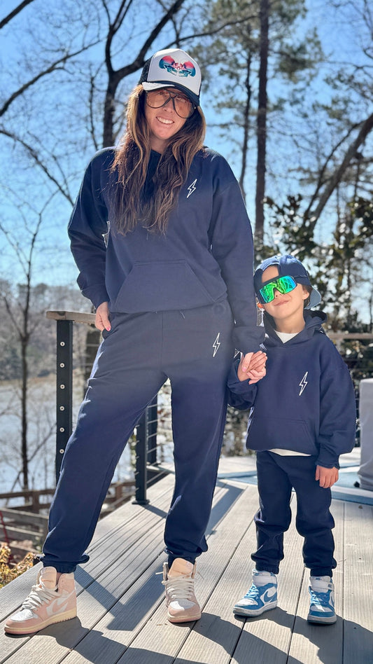 Adult and youth models wearing matching navy Big Brain Energy Hoodies and sweatpants, all with the Ladyspinedoc signature lightning bolt design. Adult model also wearing the Cranial Love Trucker Hat.
