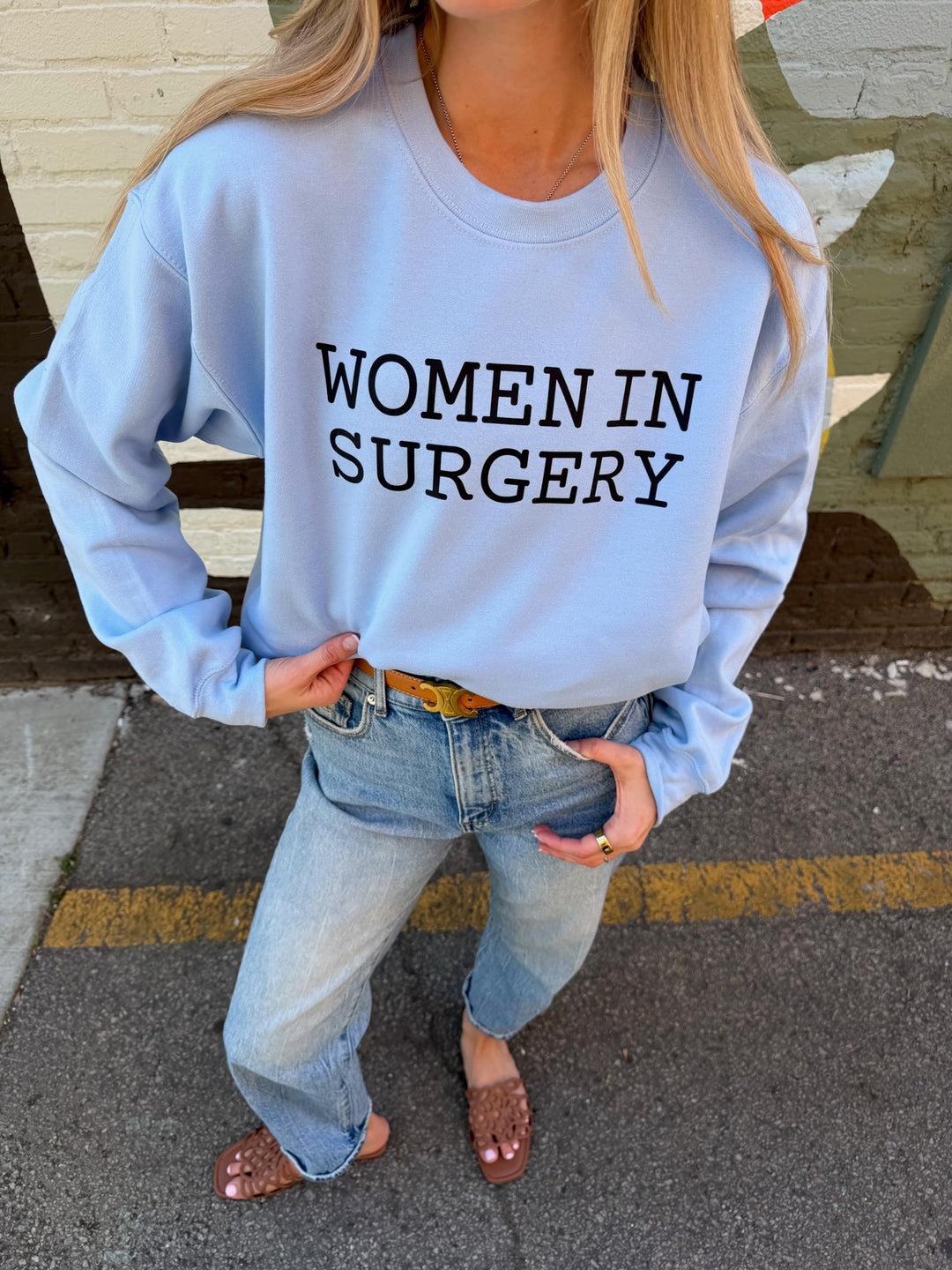 Person wearing a light blue sweatshirt with 'WOMEN IN SURGERY' text, standing on a street.