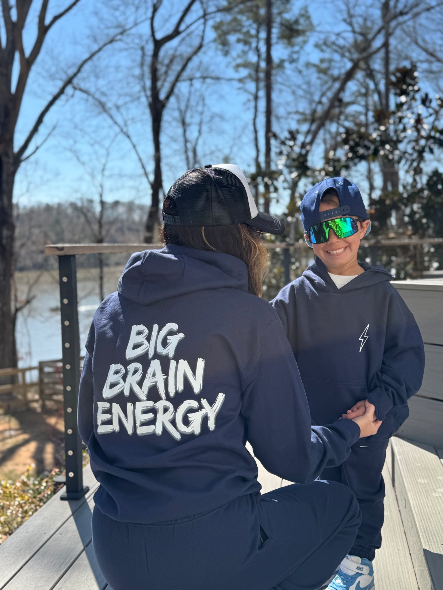 Adult model wearing Navy hoodie with "BIG BRAIN ENERGY" printed in bold white capital letters across the back. Child model showing front view of Big Brain Energy Youth Hoodie with lightning bolt on upper left chest and matching Big Brain Energy Youth Sweatpants.
