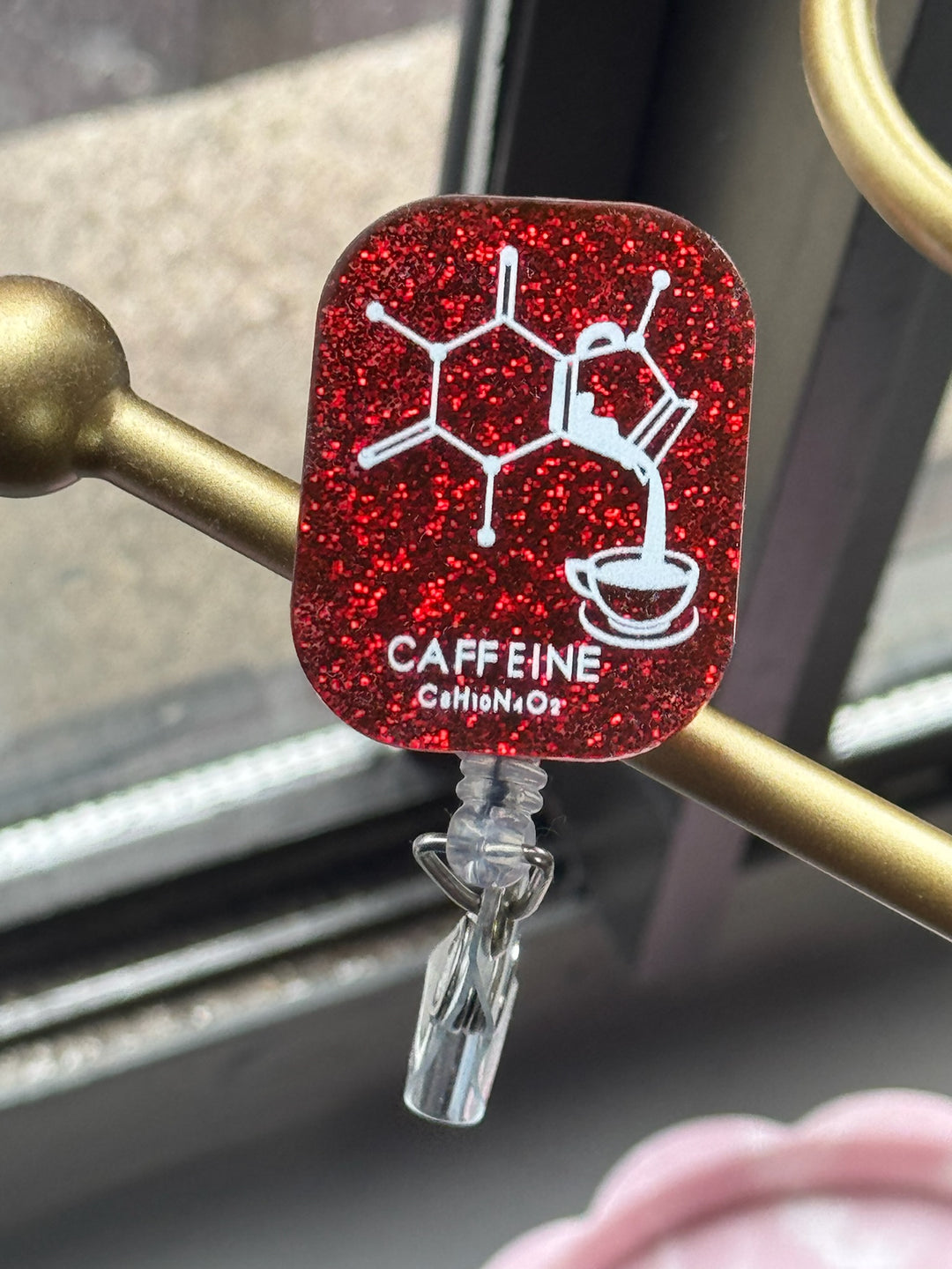 Ladyspinedoc x Dr. Beachgem Red Sparkly Enerjava Badge Reel - Retractable with Alligator Clip and Caffeine Molecule Design on a dark red glitter background, clipped to a gold stand.