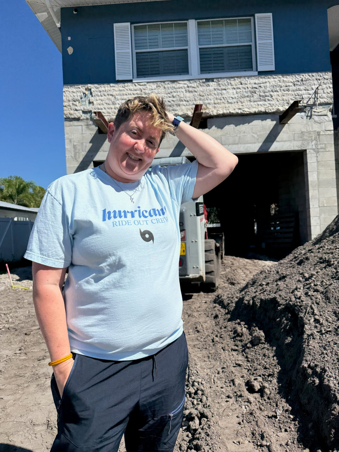 Dr. Beachgem standing in front of her house under construction with a blue Hurricane Ride Out Crew tee shirt on.