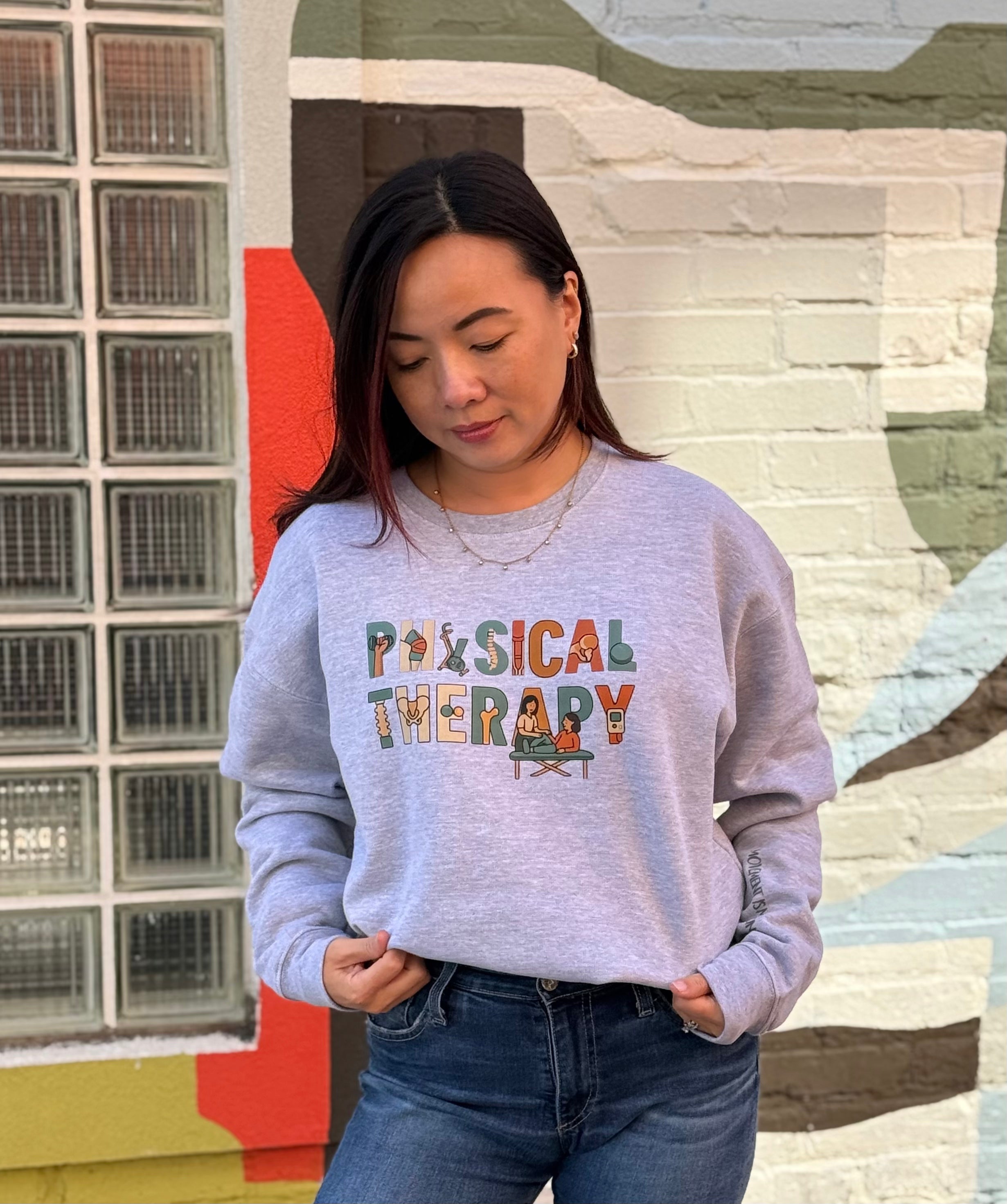 Woman wearing a heather gray sweatshirt with 'Physical Therapy' text and colorful design, standing in front of a textured wall.