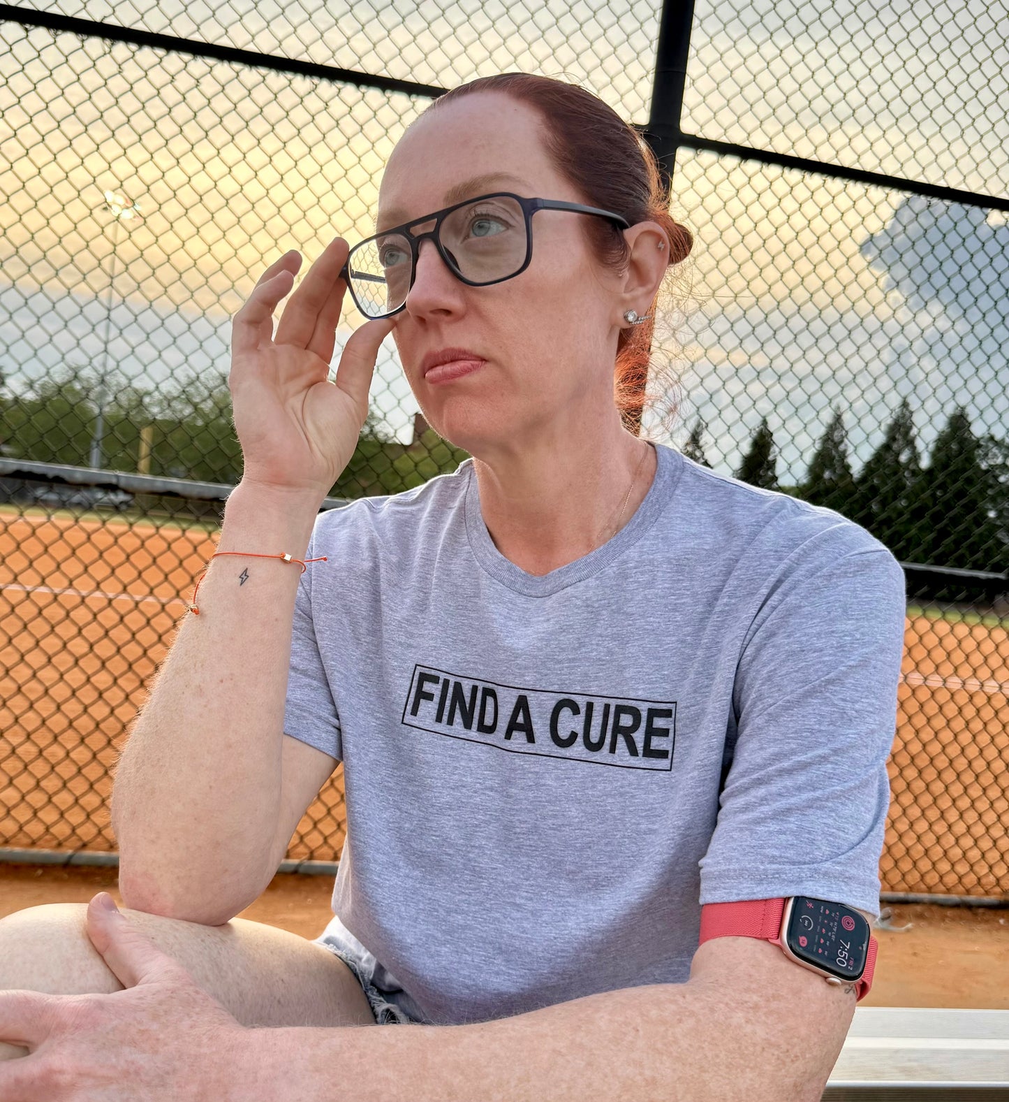 Ladyspinedoc (Dr. Betsy Grunch) sitting on bleachers in light gray LFG Brain Tumor Awareness t-shirt that reads “FIND A CURE” in black across the chest