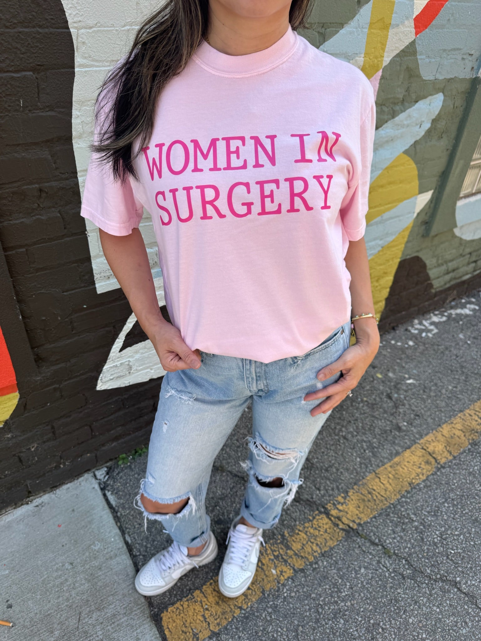 Person wearing a pink 'WOMEN IN SURGERY' t-shirt standing on a street.