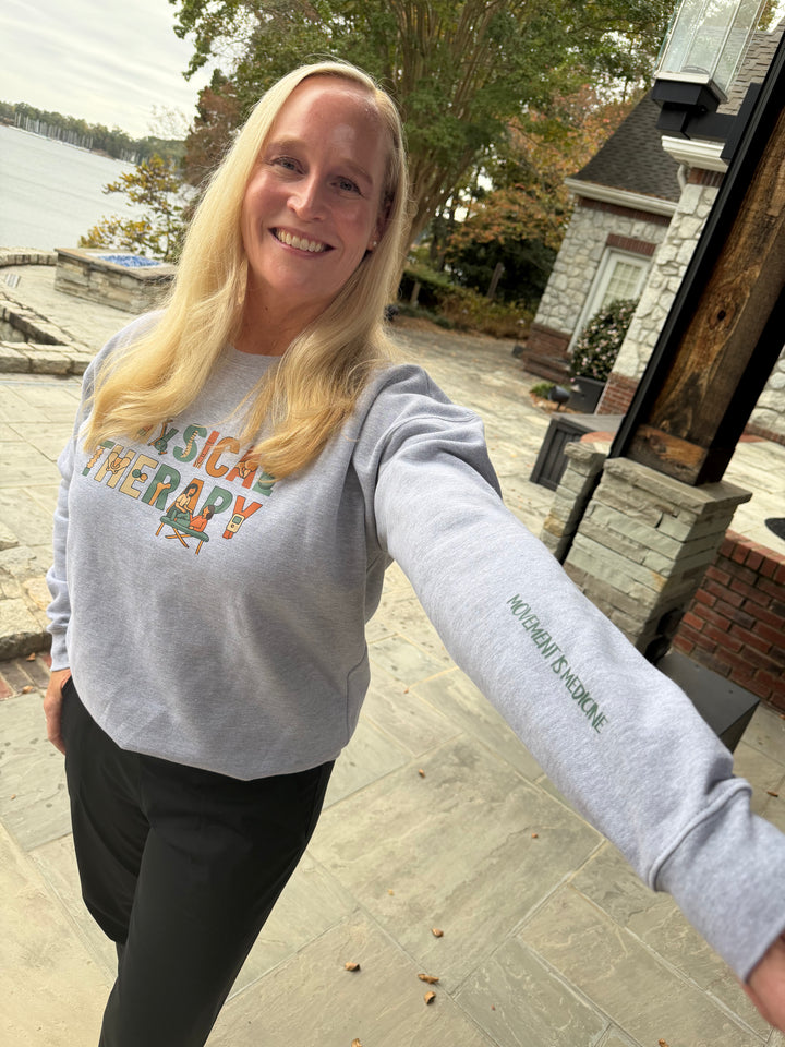 Woman wearing a gray sweatshirt with 'Physical Therapy' and 'Movement is Medicine' text, standing outdoors by a lake.