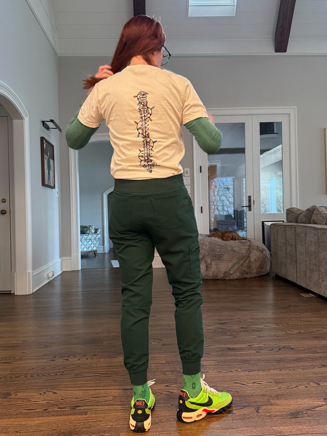 Ladyspinedoc wearing the Merry and Aligned beige t-shirt with a spine design wrapped in colorful lights, in a room with wooden flooring and walls.