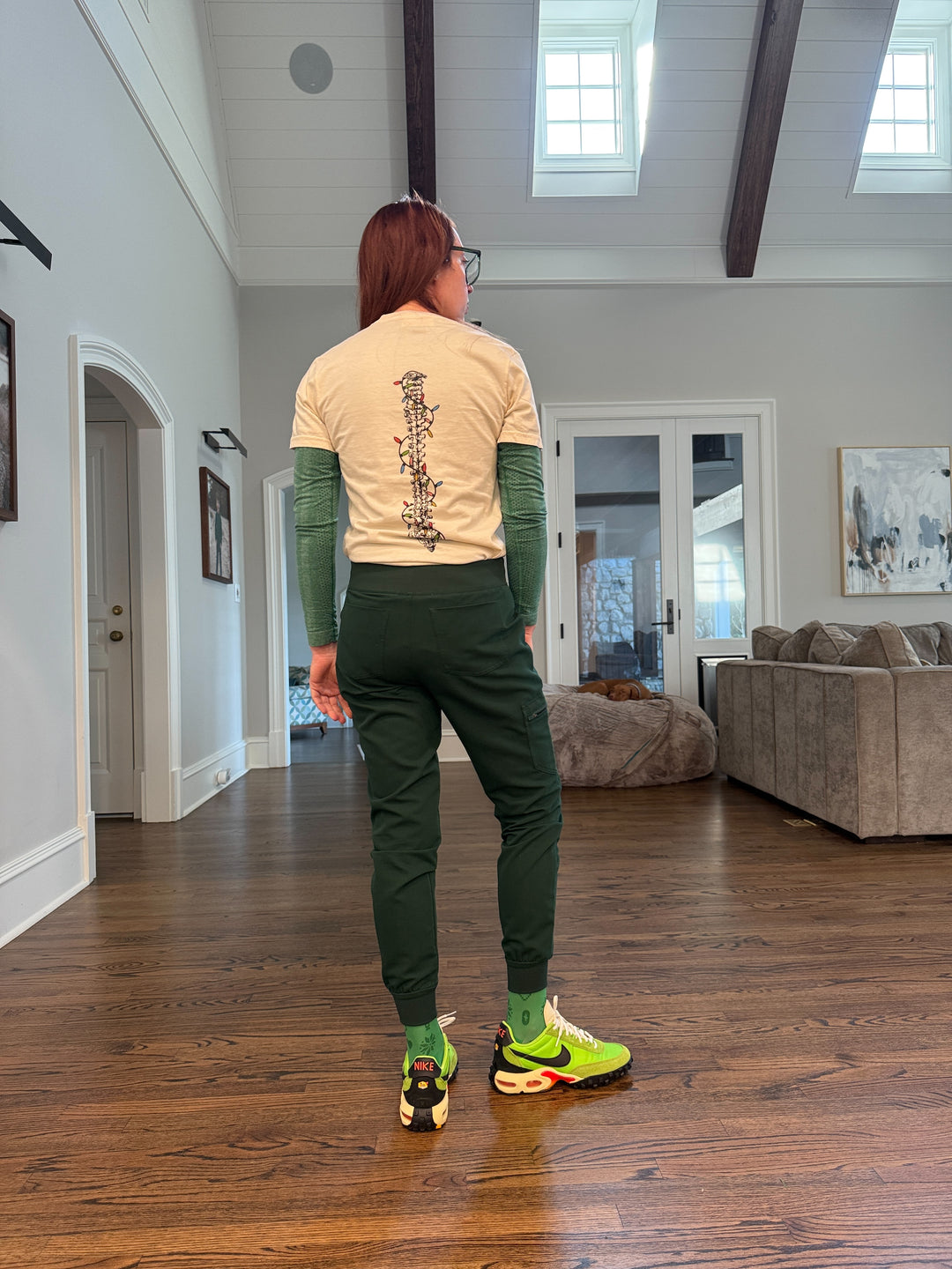 Ladyspinedoc wearing a beige t-shirt with a spine design wrapped in colorful lights, in a room with wooden flooring and walls.