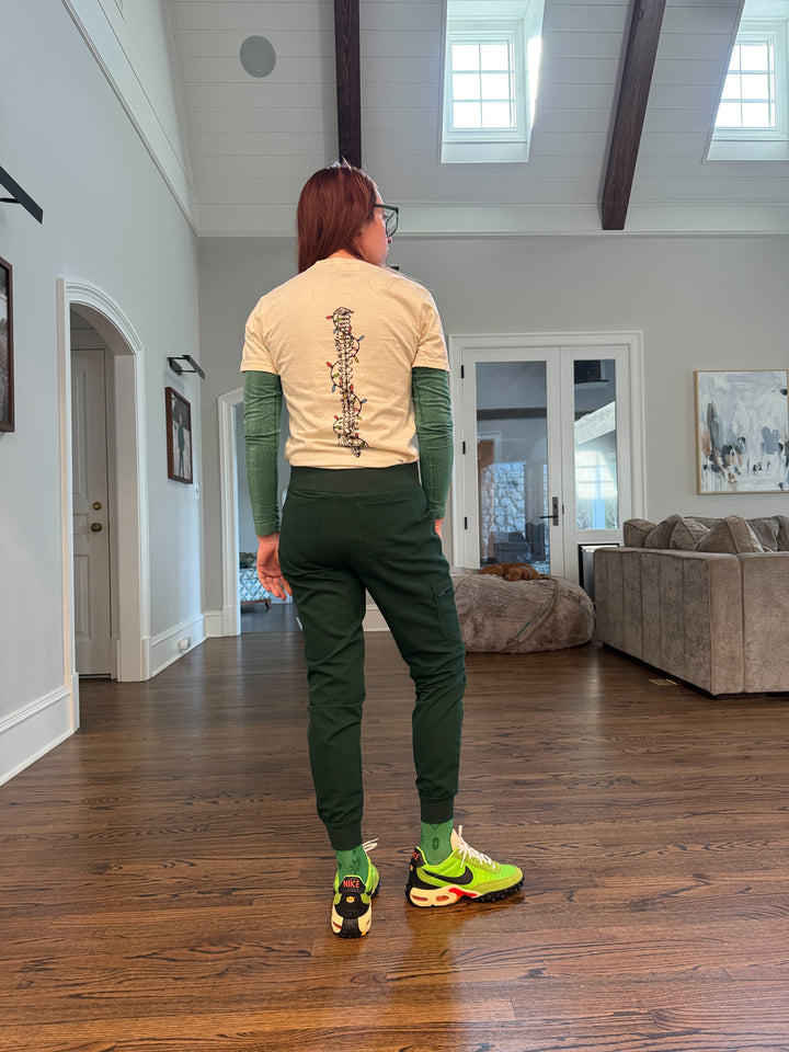 Ladyspinedoc wearing a beige t-shirt with a spine design wrapped in colorful lights, in a room with wooden flooring and walls.