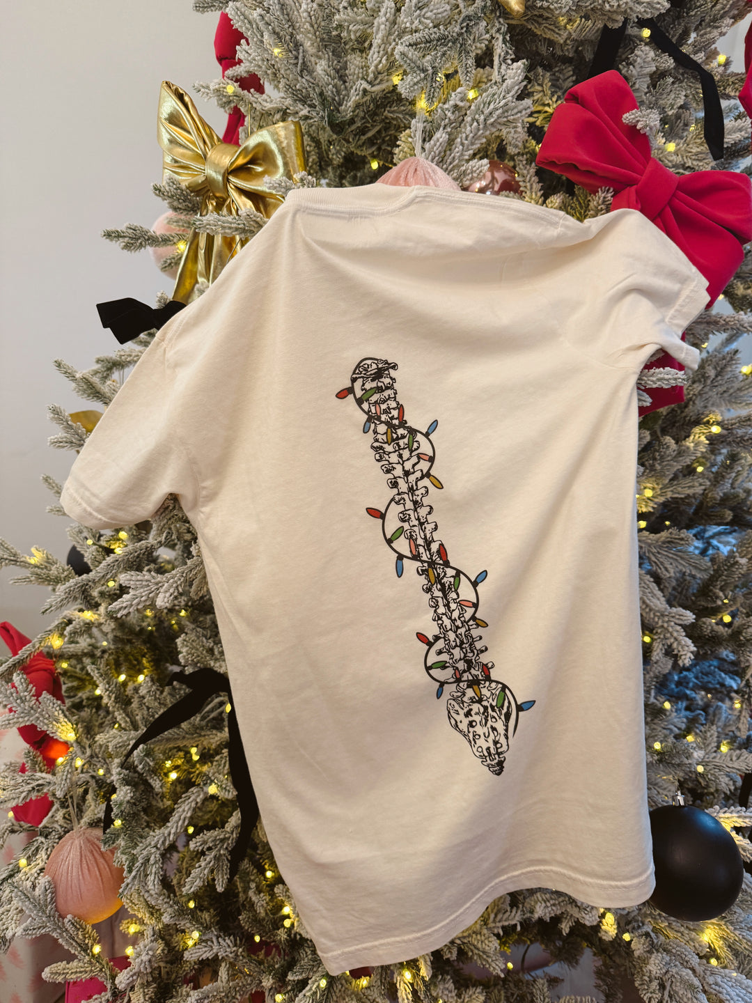 The Merry & Aligned beige t-shirt with a back spine design wrapped in colorful lights hanging on a Christmas tree
