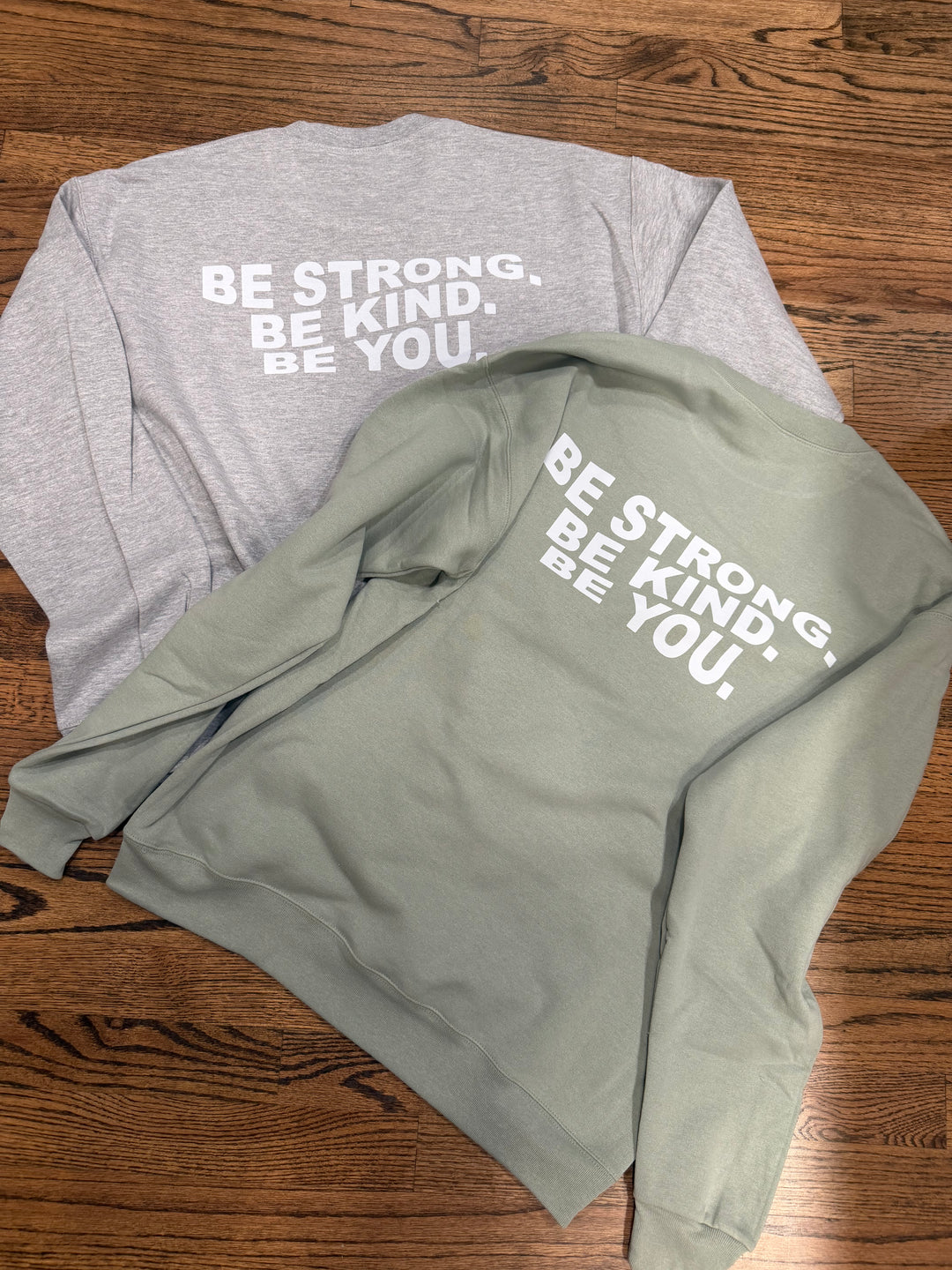 Two Ladyspinedoc Be You sweatshirts, one gray and one green, with motivational text on a wooden surface.
