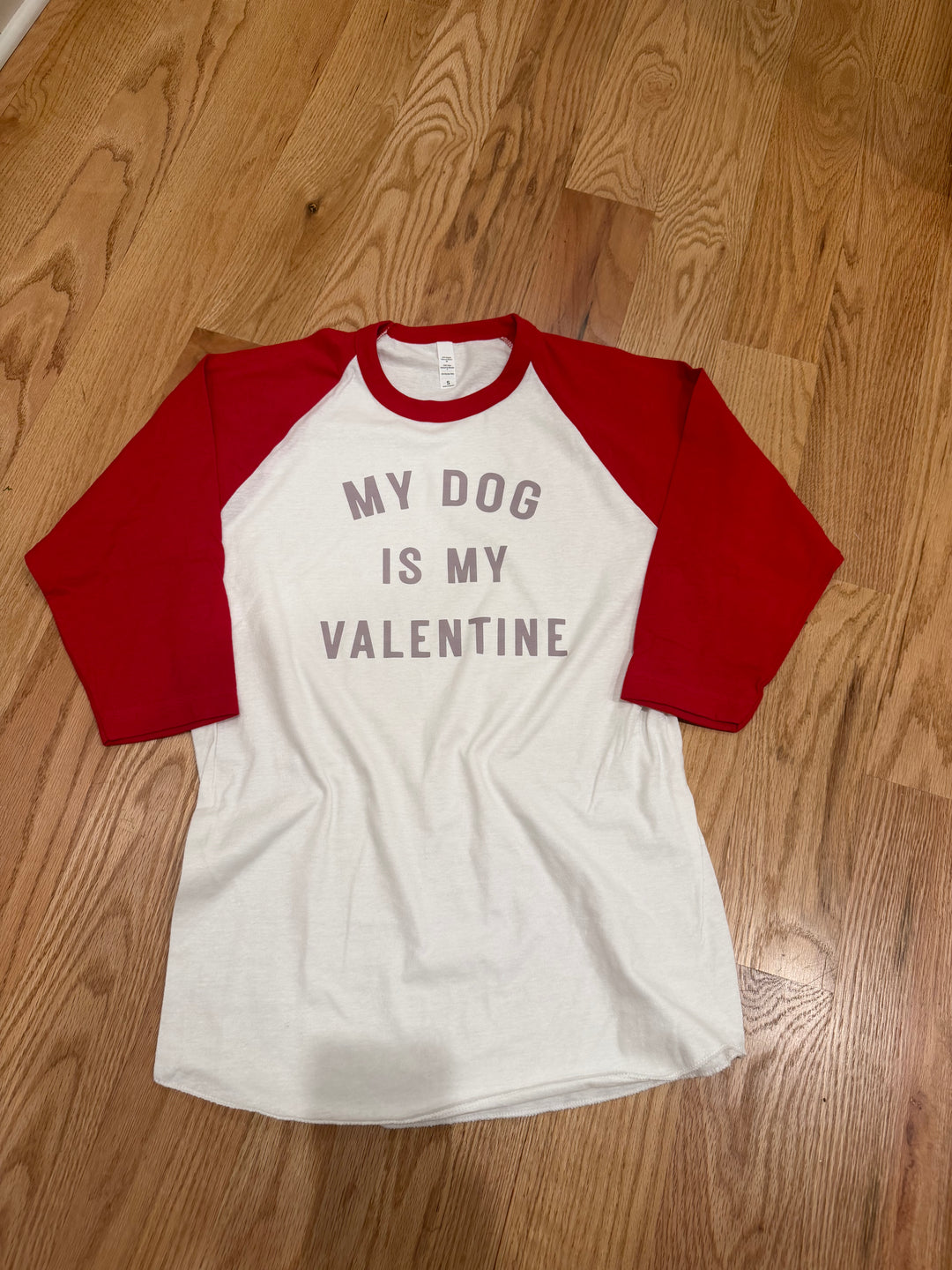 White and red baseball shirt with 'My dog is my valentine' text on a wooden floor.