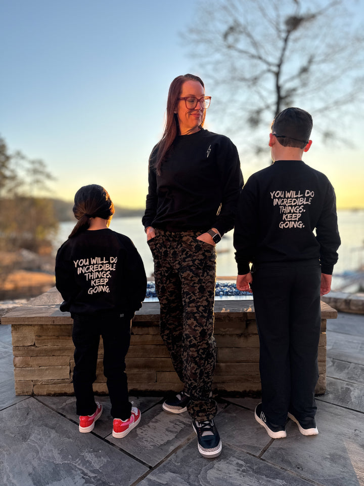 Dr. Betsy Grunch (Ladyspinedoc) looking down toward the male youth model and showing the front of the black Incredible Sweatshirt. Youth models on either side of Dr. Grunch are facing backward, wearing the black youth versions of the Incredible Sweatshirt and showing the bold white brushstroke message: "YOU WILL DO INCREDIBLE THINGS. KEEP GOING." on the back.
