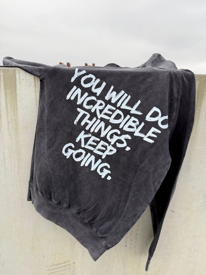 The dark gray Incredible Hoodie laying over the top of a concrete wall showing the bold white brushstroke message: “YOU WILL DO INCREDIBLE THINGS. KEEP GOING.” back design.
