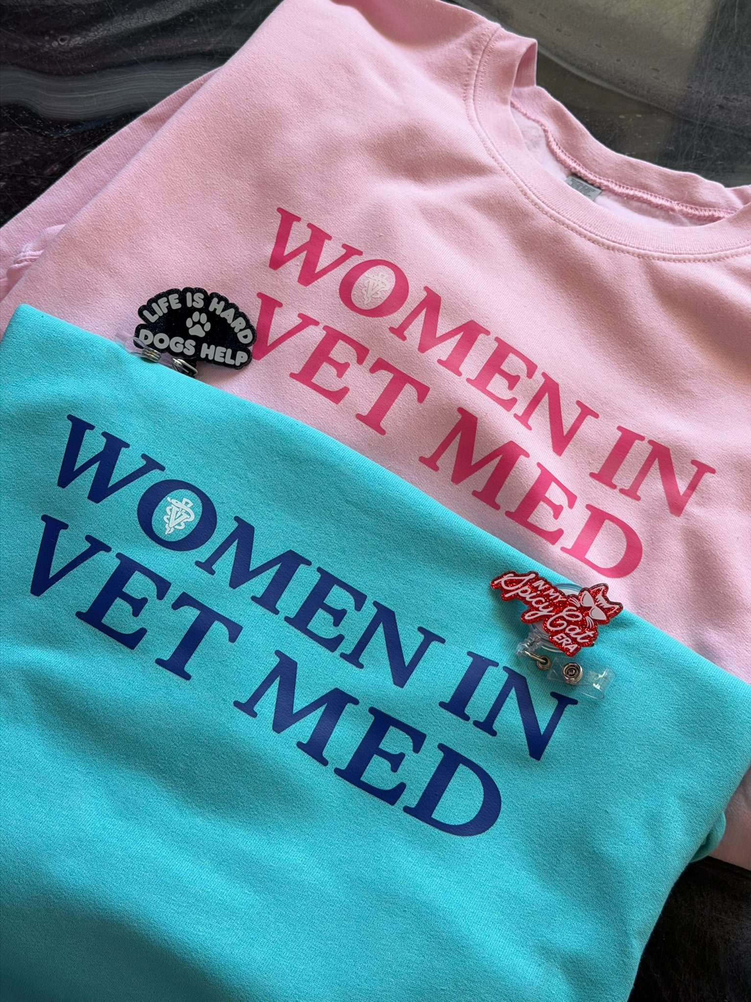Two shirts with 'Women in Vet Med' text, one pink and one teal, on a dark surface.