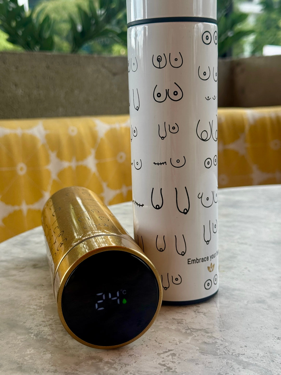 White Bosom Smart Water Bottle with Breast Design standing up on table and LED Temperature Display shown on gold version laying on its side