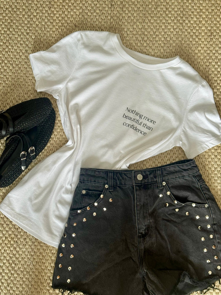 Flat lay of The Confidence Doc Confidence Tee Shirt that says "Nothing more beautiful than confidence" with black shorts and shoes