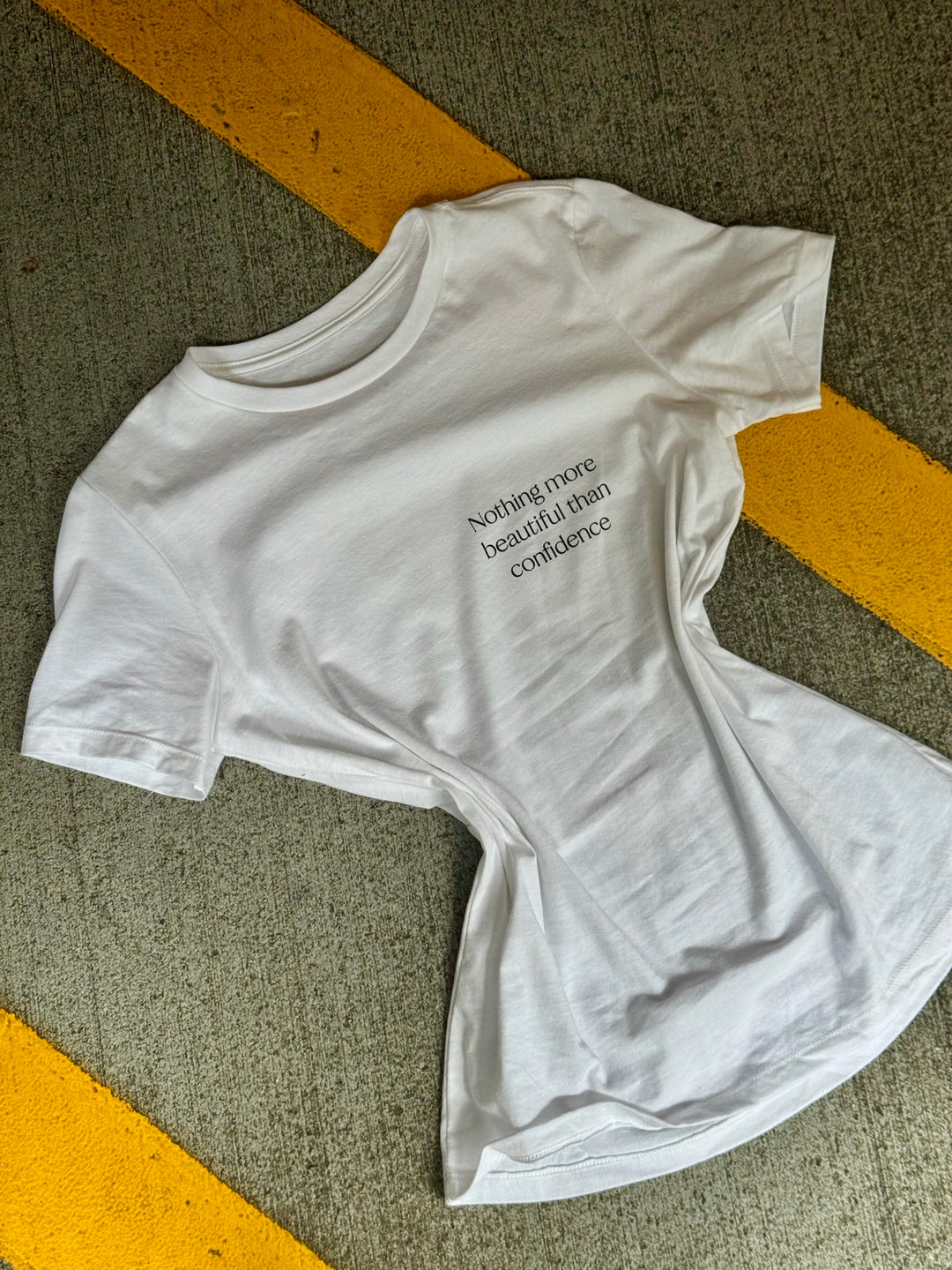 Flat lay of The Confidence Doc Confidence Tee Shirt on pavement with "Nothing more beautiful than confidence" motto