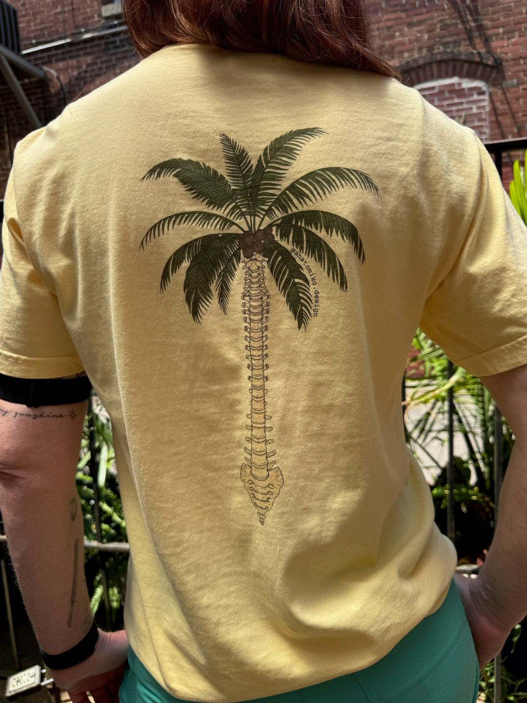 Ladyspinedoc wearing a yellow t-shirt with a spine palm tree design on the back, standing outdoors.