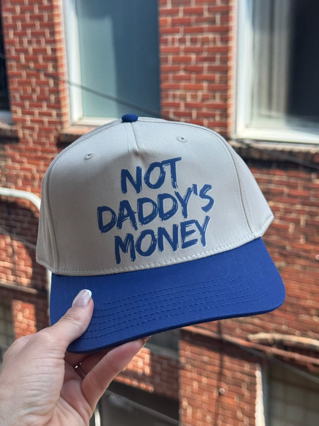 Beige and blue cap with 'Not Daddy's Money' text held in front of a brick building.