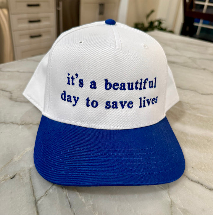 White cap with blue embroidered "it's a beautiful day to save lives" with blue brim laying on a marble surface