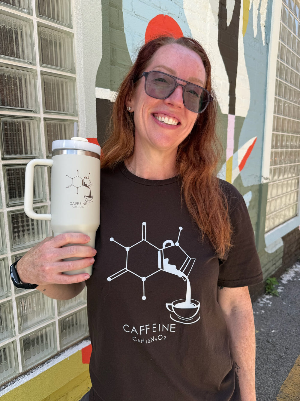 Ladyspinedoc wearing a brown Enerjava  t-shirt with a caffeine molecule design, holding a white tumbler, in front of a colorful mural.