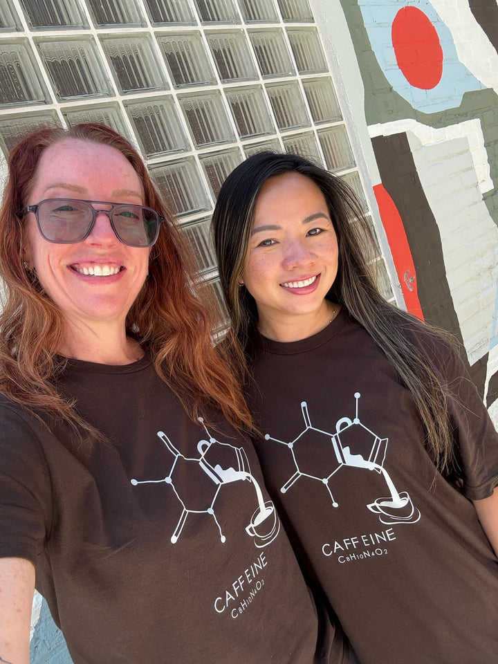 Ladyspinedoc and Jenn wearing brown Enerjava t-shirts with a molecule design and 'CAFFEINE' text, standing in front of a colorful wall.